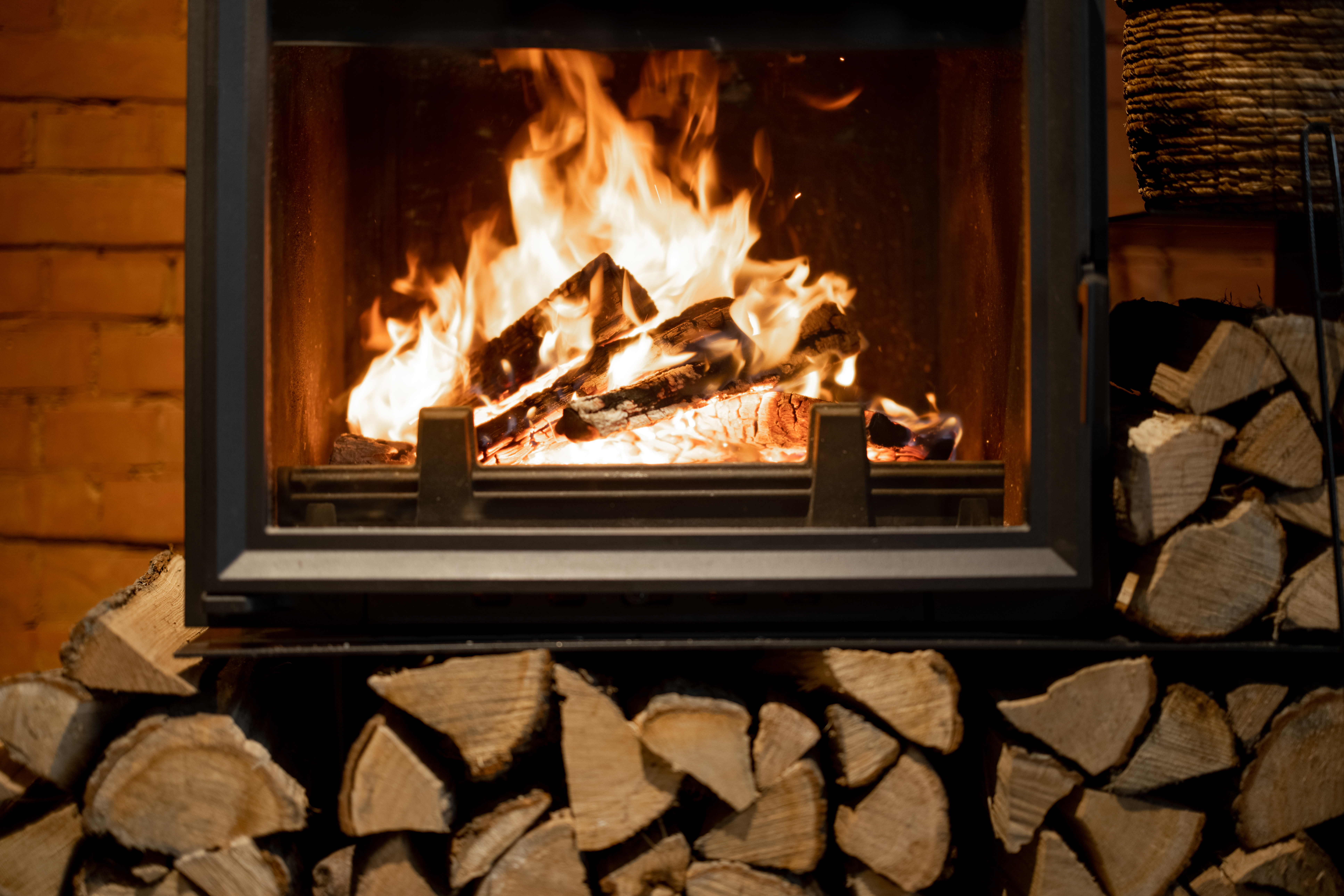 A close-up of a wood burner stove with a warm fire burning inside, surrounded by a neat pile of split firewood.