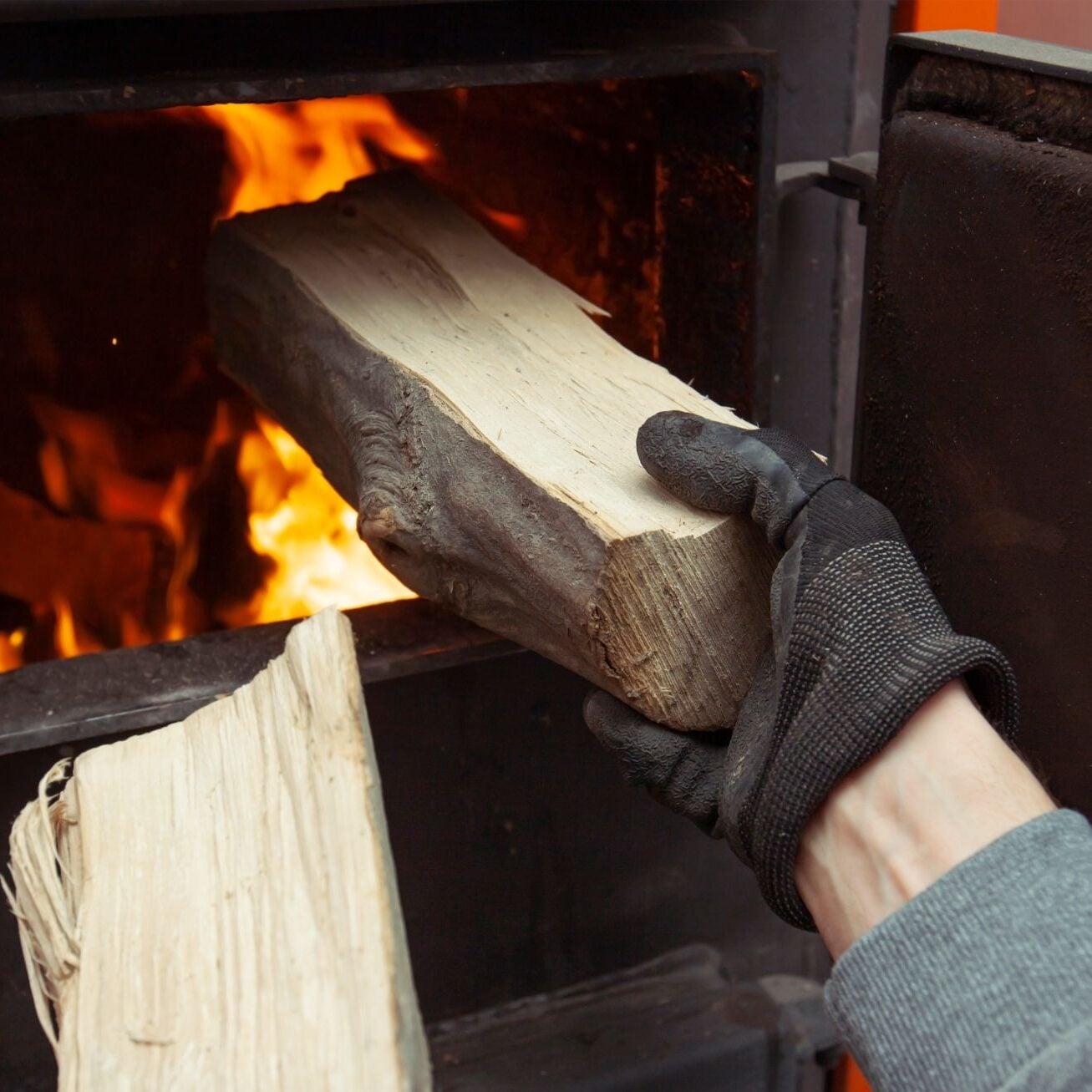 A gloved hand adding a log to a burning wood stove. The fire is burning brightly in the background.